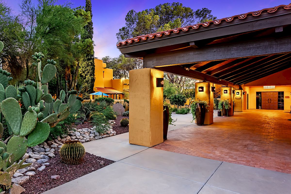 The image shows a Southwestern-style building with a tile roof, a covered entrance, and cactus plants in the foreground, surrounded by desert landscaping.
