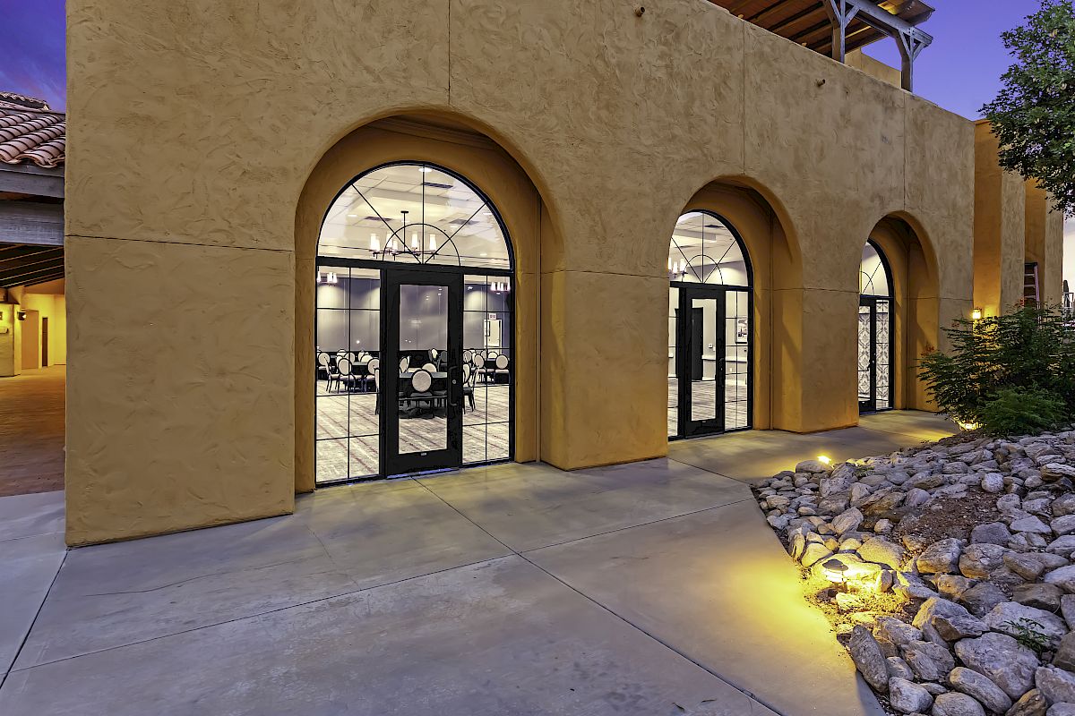 The image shows a building with three large arched windows and glass doors, with a well-lit interior and landscaped rocks outside.