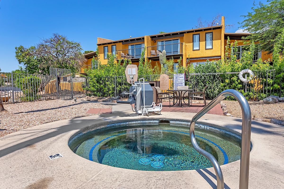 Image of a hot tub in outdoor setting with handrails, a pool lift, and surrounded by greenery and residential buildings in the background.