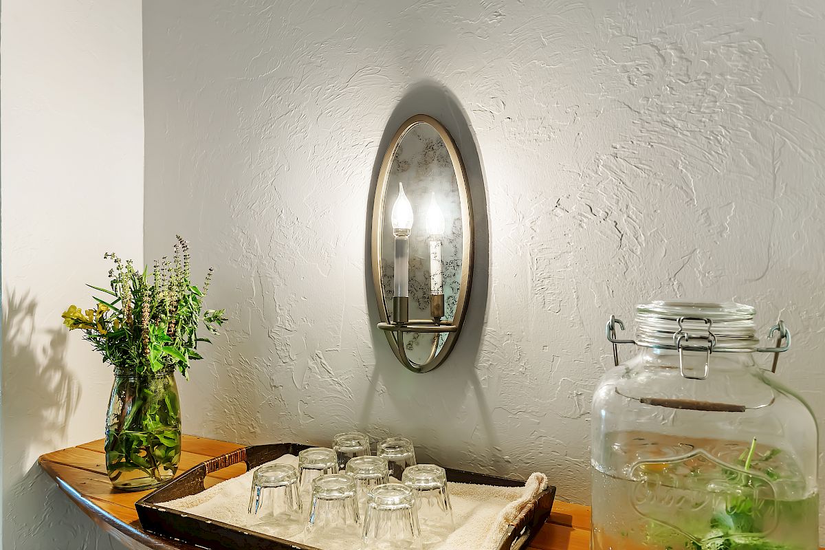A mirrored wall light, a vase with flowers on a wooden shelf, a tray with glasses, and a glass dispenser with infused water and herbs.