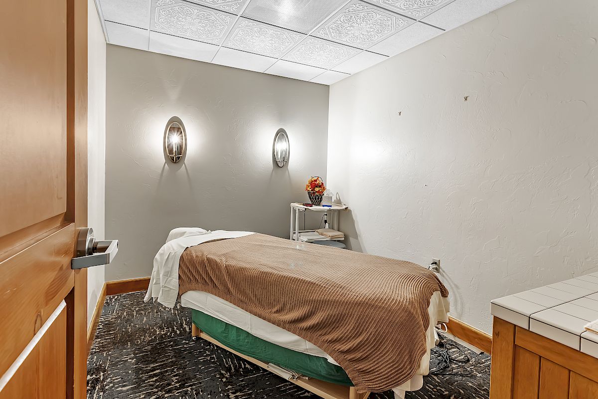 This image shows a small massage room with a massage table, wall lights, a small shelf with few items, and a calm ambiance.