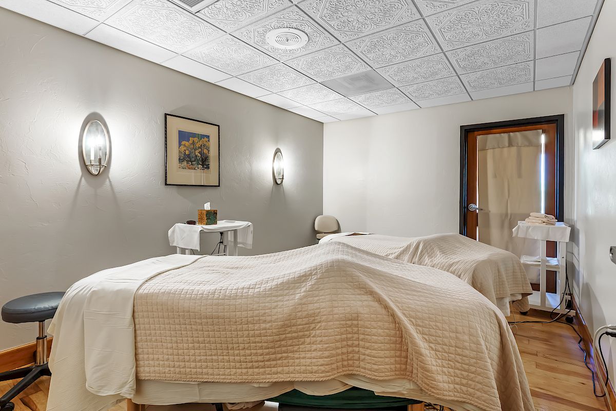A peaceful spa room with two massage tables, covered with cream-colored blankets, soft lighting, and minimal decor is shown.