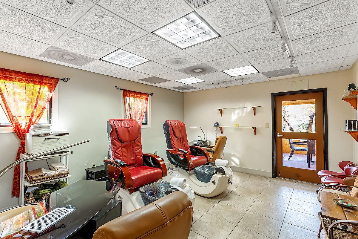 A brightly lit salon room with two pedicure chairs, an office chair, shelves, windows with curtains, and a door leading outside.