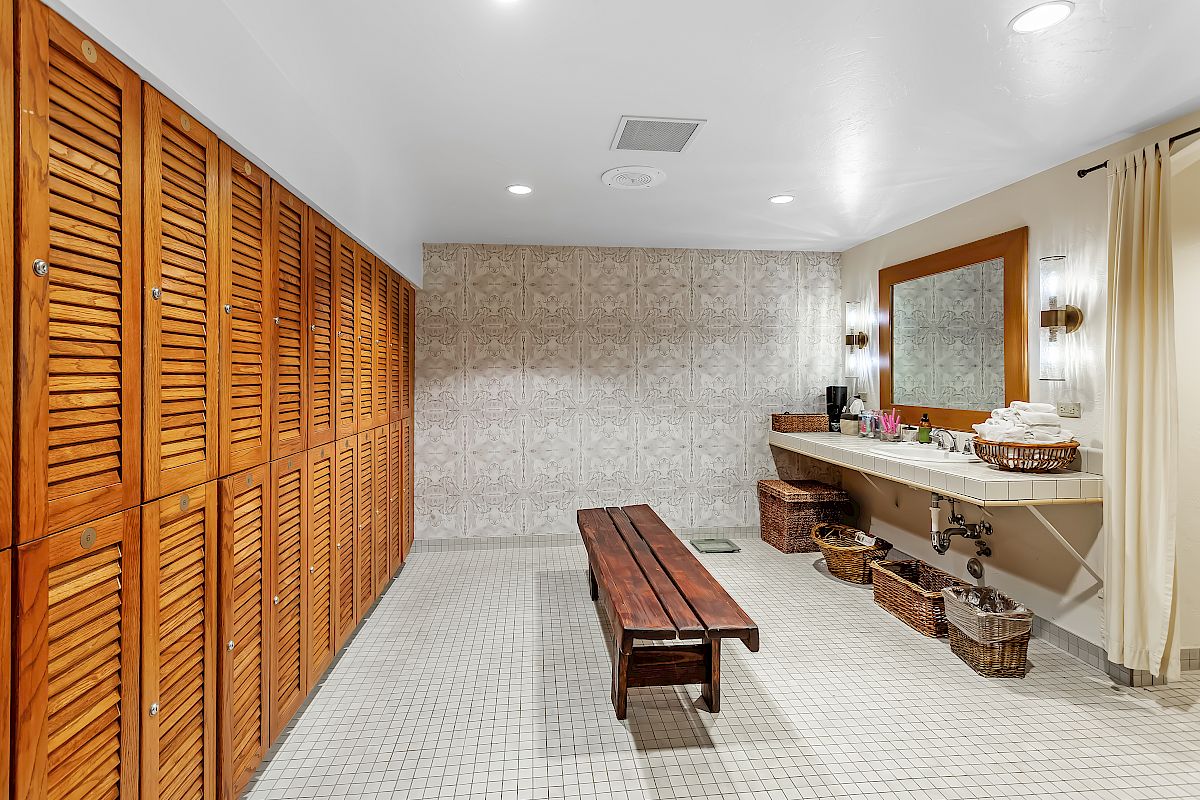 The image shows a spacious locker room with wooden lockers, a bench in the center, and a vanity area with sinks, mirrors, and wicker baskets.