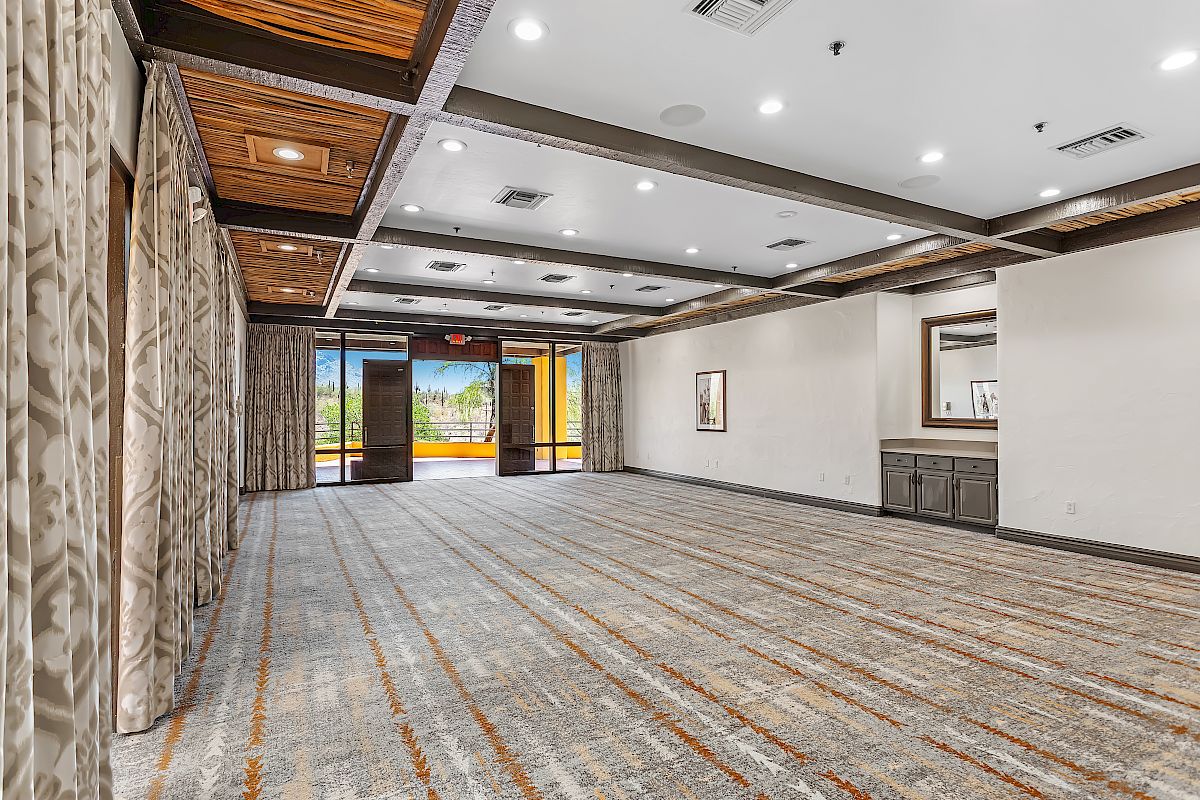 An empty, modern conference room with a patterned carpet, wood-paneled ceiling, large windows, and an open outdoor view through glass doors, ending the sentence.