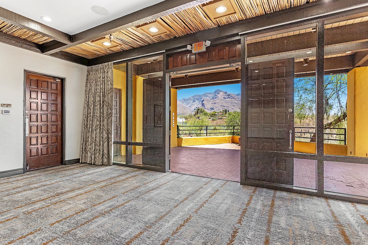 A spacious room with patterned carpeting, wooden ceiling beams, large glass doors opening to a balcony with mountain views beyond.