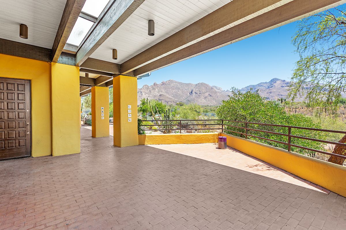 A covered outdoor corridor with yellow pillars, brick flooring, and a scenic mountain view in the distance on a clear, sunny day.