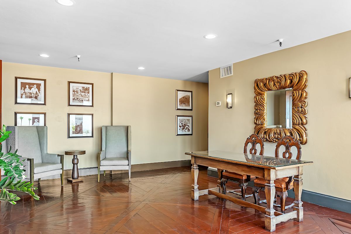 The image shows an indoor space with a wooden desk, ornate mirror, chairs, a small table, and framed pictures on the walls.