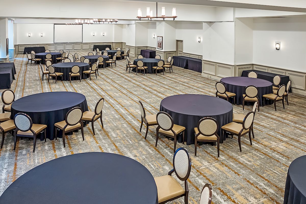 The image shows a banquet hall with round tables covered in black tablecloths and surrounded by chairs, the floor is carpeted, and lighting fixtures hang from the ceiling.