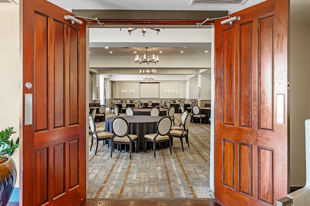 An open door reveals a spacious, elegant conference room set with round tables and chairs, ready for a meeting or event, with chandeliers overhead.