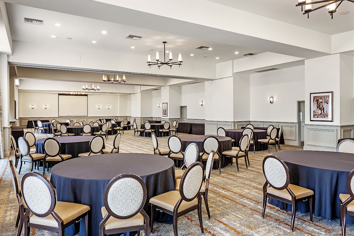 The image shows an empty banquet hall with round tables covered in black tablecloths and beige cushioned chairs, under modern chandeliers.