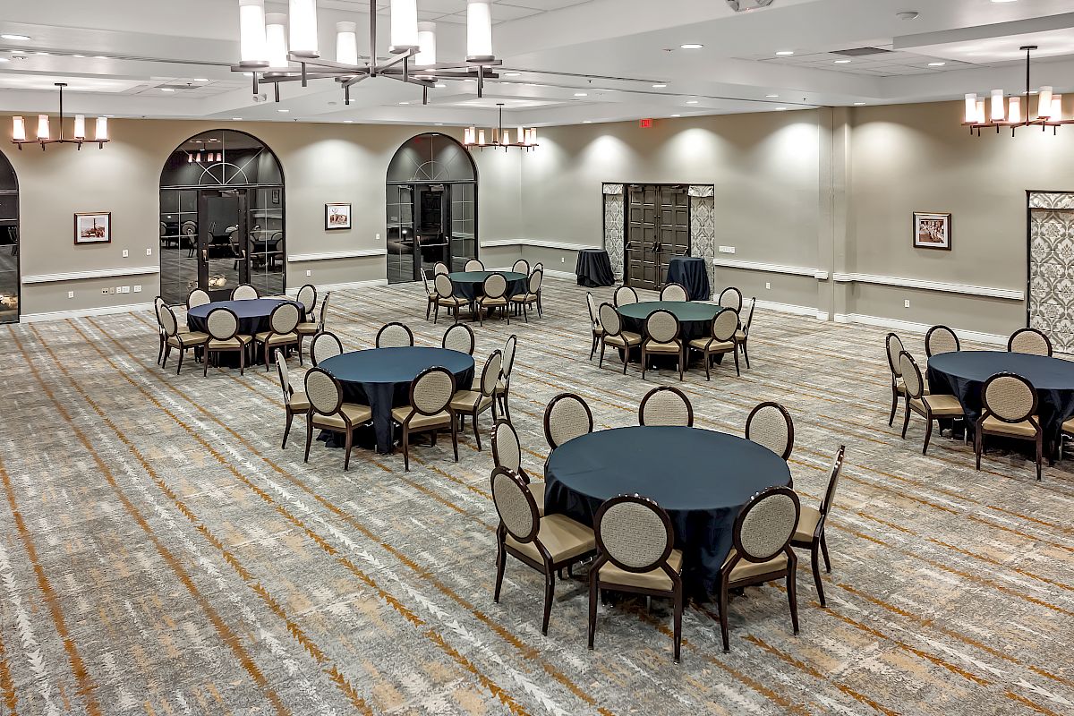 The image shows a luxurious banquet hall with round tables covered in dark tablecloths, chairs, chandeliers, artwork, and patterned carpet.