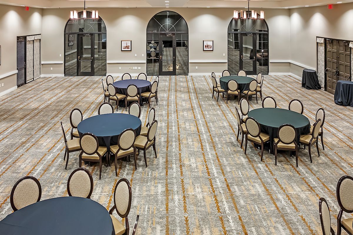 An empty, well-lit banquet hall with round tables and chairs arranged evenly across a carpeted floor. It features large windows and elegant lighting fixtures.