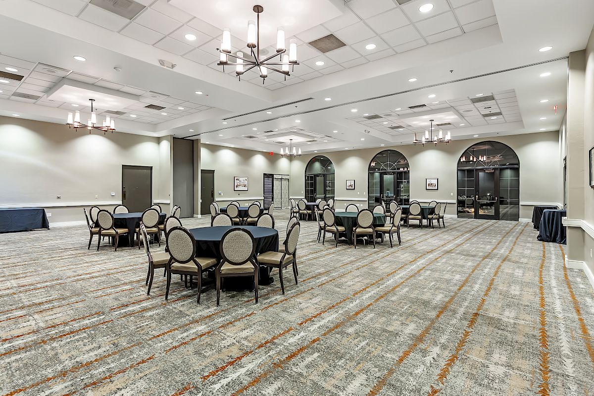 The image features an empty, well-lit banquet hall with round tables and chairs arranged sparsely, chandeliers on the ceiling, and large arched windows.