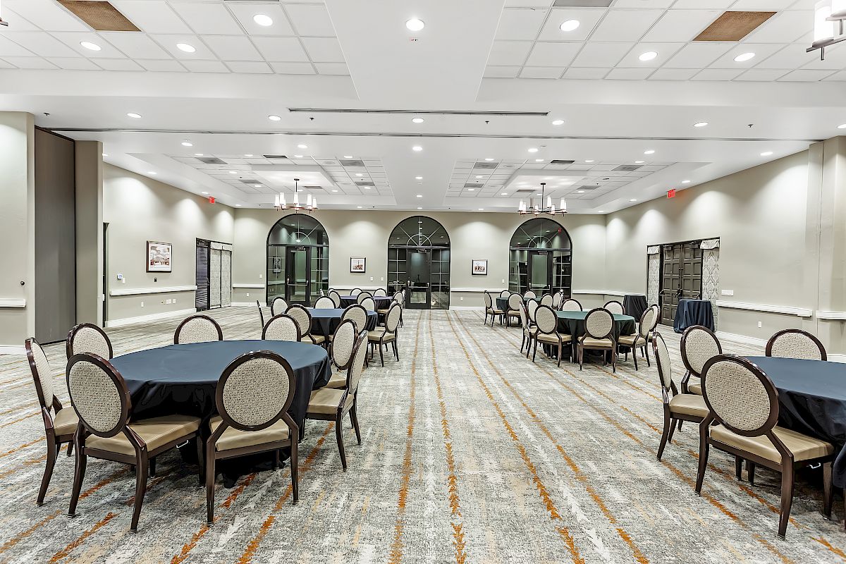 A spacious, well-lit conference room with circular tables and chairs arranged in rows, featuring large windows and elegant chandeliers with wall decor.