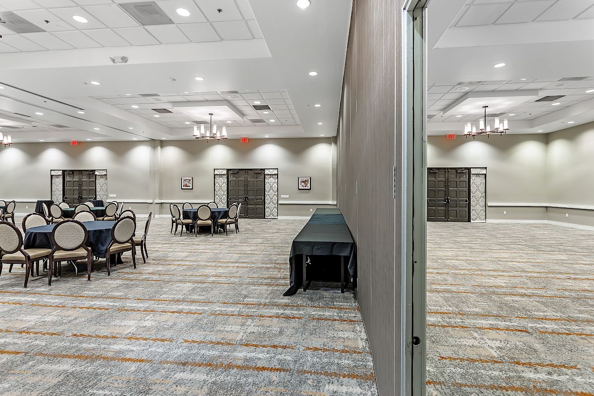 A spacious banquet hall with round tables and chairs, partitioned into two separate areas, with chandeliers on the ceiling and light-colored carpeting.