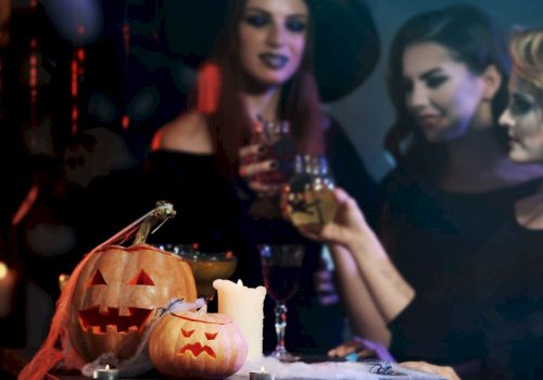 Three people in Halloween costumes holding drinks, with two carved pumpkins and a candle in the foreground on a dimly lit table.