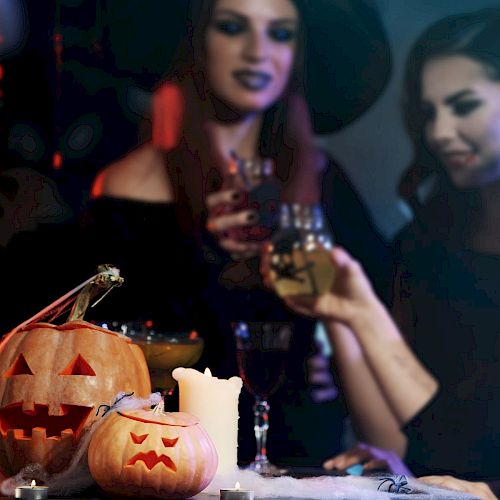 Three people in Halloween costumes holding drinks, with two carved pumpkins and a candle in the foreground on a dimly lit table.