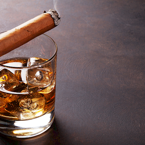 A glass of whiskey with ice and a partially smoked cigar rests on top, set against a dark background.