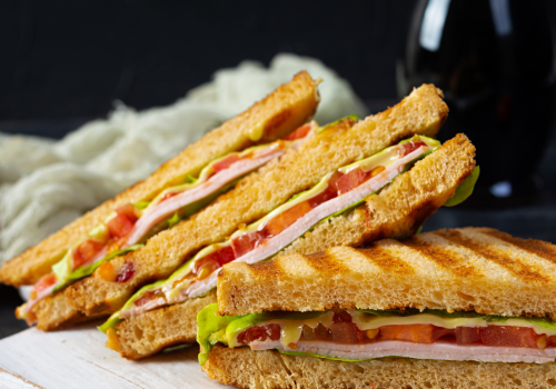 The image shows grilled sandwiches with layers of ham, cheese, lettuce, and tomato, cut into triangles and stacked.