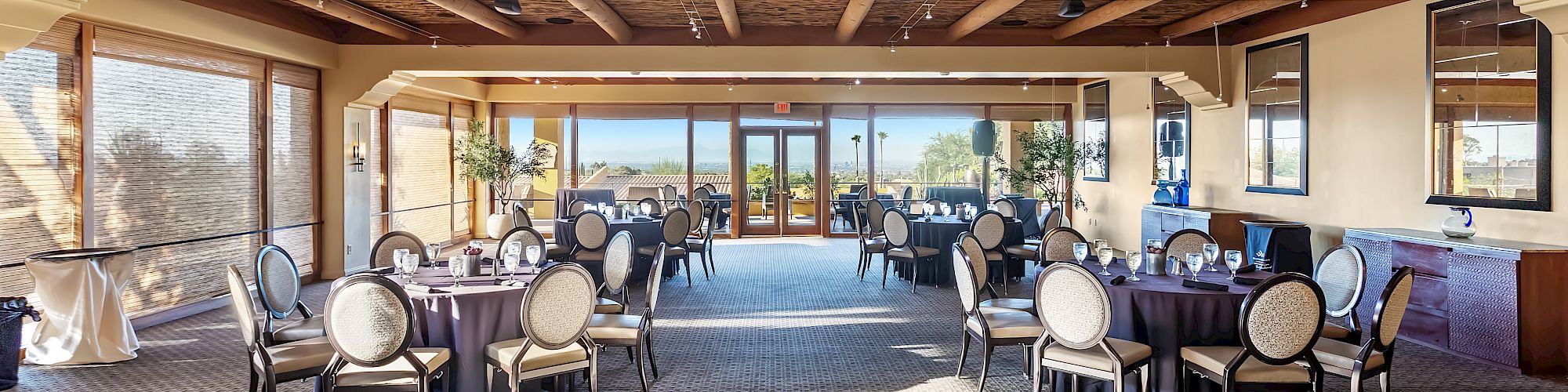 A spacious banquet or dining area with round tables and chairs, large windows, and a view of an outdoor area with greenery and blue sky.