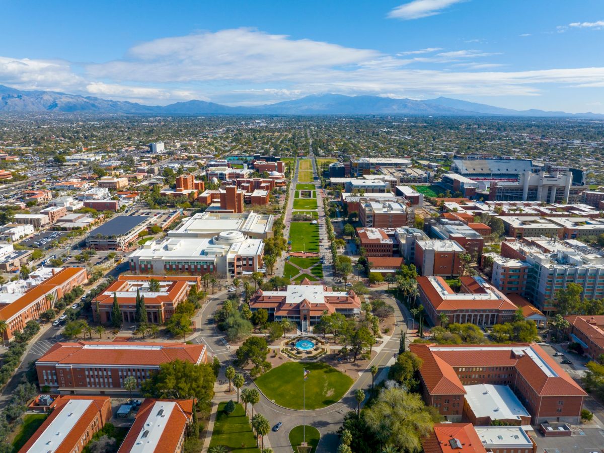 Aerial view of a sprawling campus with red-roofed buildings, green spaces, roads, and distant mountains under a blue sky.