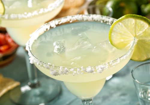 A margarita cocktail with ice and a lime garnish, served in a glass with a salted rim, is shown in the image.