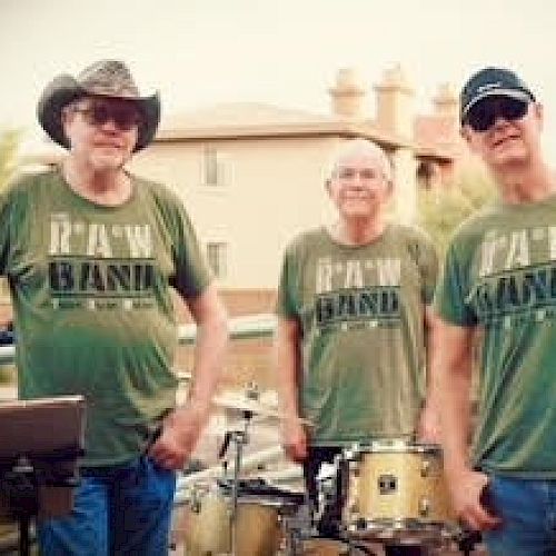 Three men in matching RAW BAND shirts stand outdoors near musical equipment, including a drum set, with greenery in the background.