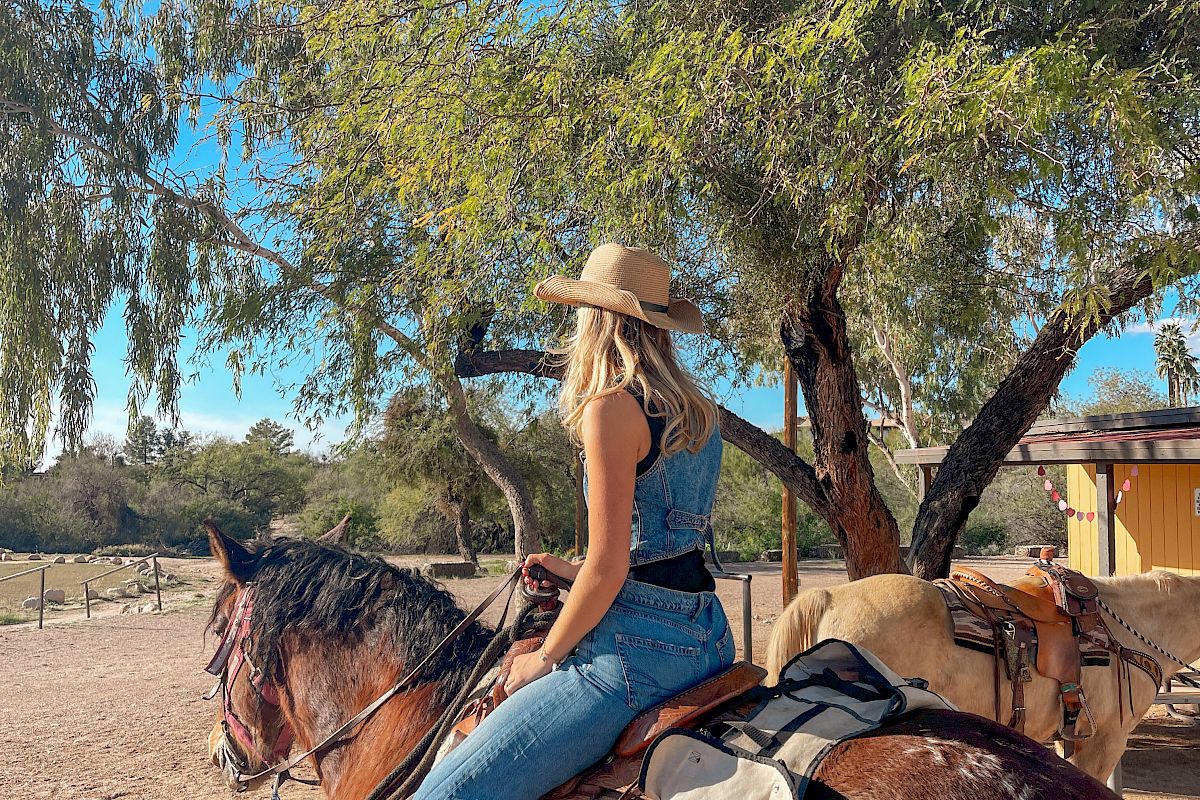Woman in a straw hat sits on a saddle horse in a sunny ranch, with big trees overhead.