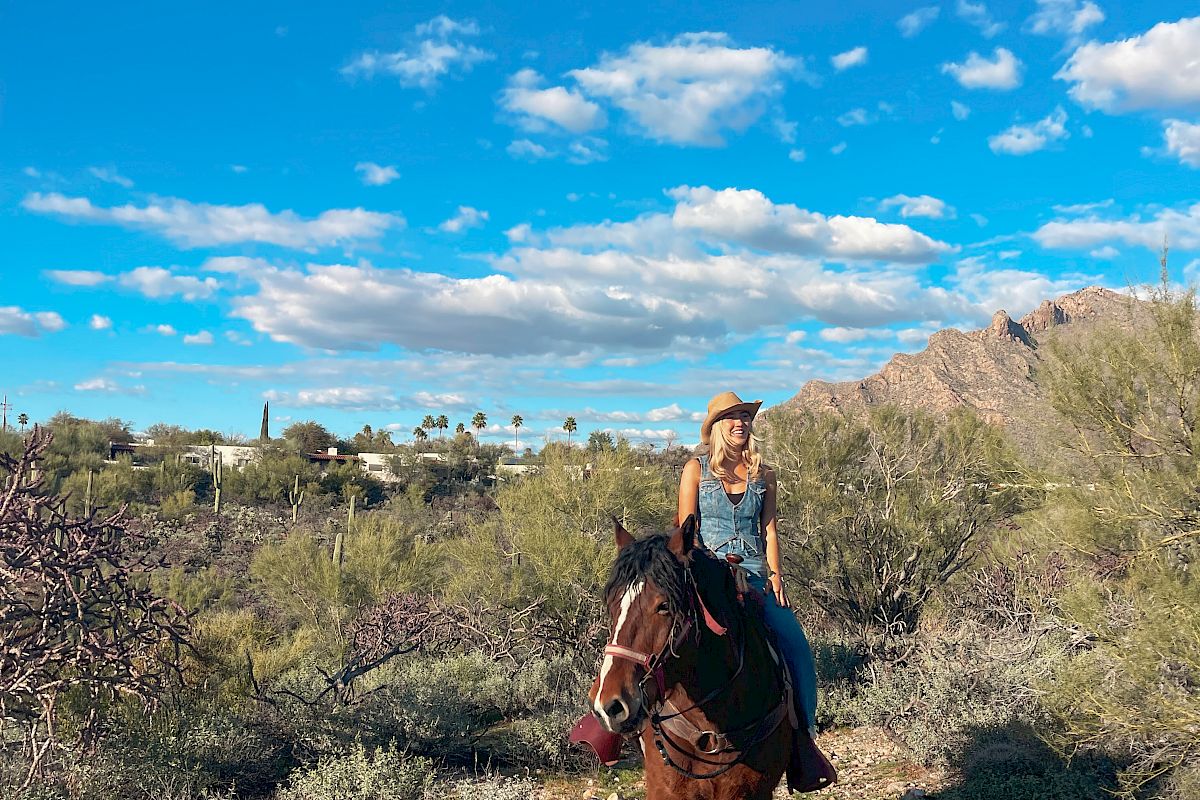 Woman riding a horse through a desert landscape with cacti, mountains, and a bright blue sky full of clouds.